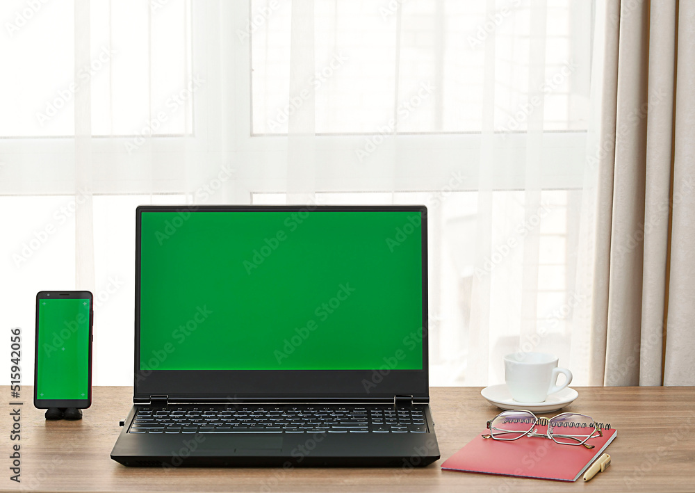 Fototapeta premium Laptop and mobile phone with green screen on wooden table against window background