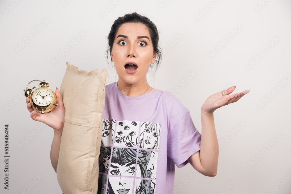 Obraz premium Shocked woman holding alarm clock and cushion on white background