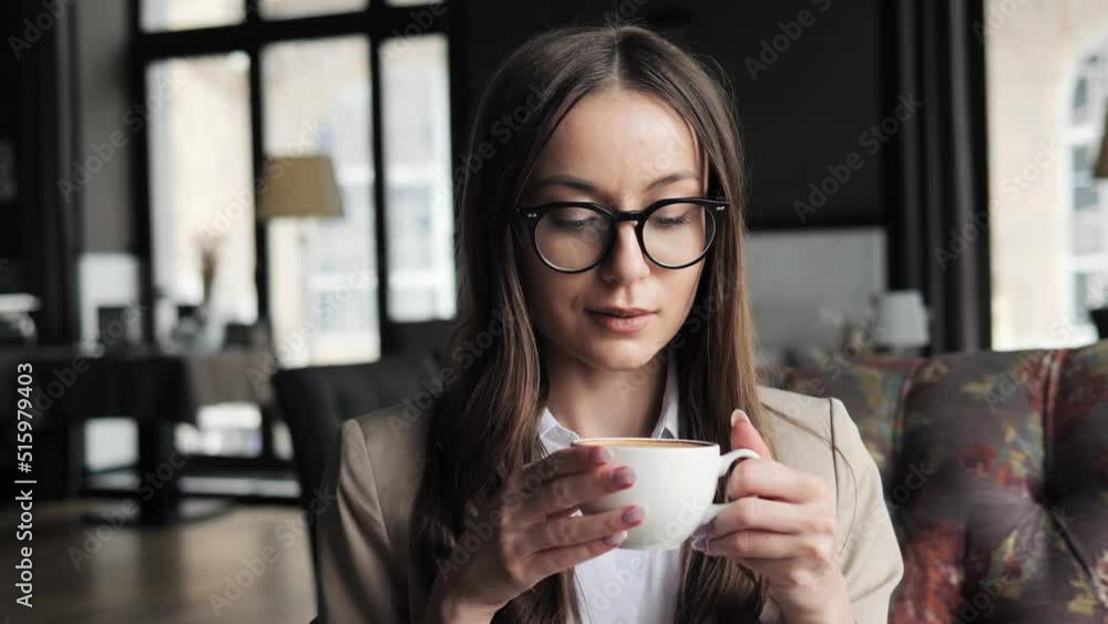 Pretty confident business woman in formal suit and eyeglasses drinking coffee in hotel lounge. Businesswoman looking posing to camera holding white cup of tea . Businesswoman stands in restaurant. Pretty confident business woman in formal suit and eyeglasses drinking coffee in hotel lounge. Businesswoman looking posing to camera holding white cup of tea . Businesswoman stands in restaurant.