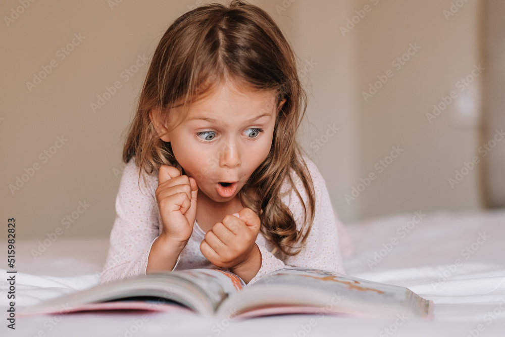 Fototapeta premium Emotional child girl read interesting book in bed. Small kid enjoy reading. Fantasy and fantastic. Developing child fantasy and imagination. Imaginary world. Fairy tale. Bedtime reading