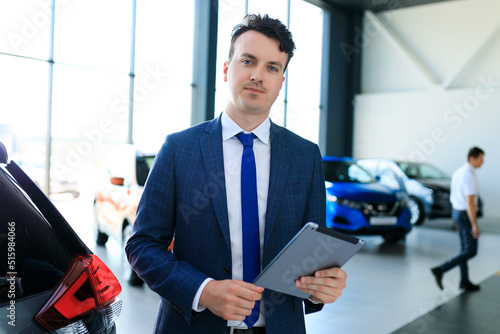A portrait of a dealership employee in a suit and tie stands in a tablet against the background of a showroom of new cars