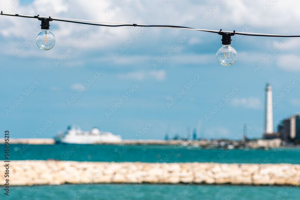 Vintage light bulbs with blue sky, white clouds, blue sea and breakwater, Bari port or harbor with cruise ship or ferry boat and lighthouse on blurred background