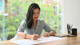 Focused young businesswoman checking financial report, project statistics, reading correspondence at wooden office desk