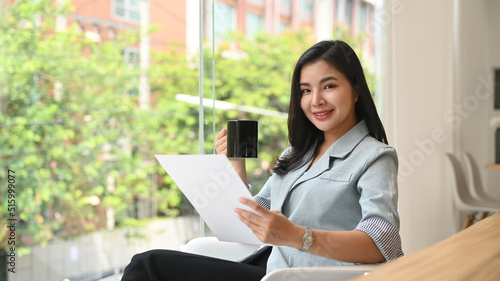 Elegant young professional woman drinking coffee and reading document in bright modern office 