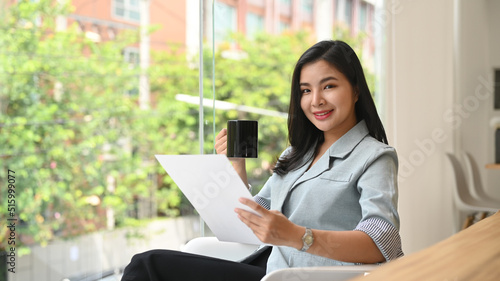 Elegant young professional woman drinking coffee and reading document in bright modern office  © Prathankarnpap