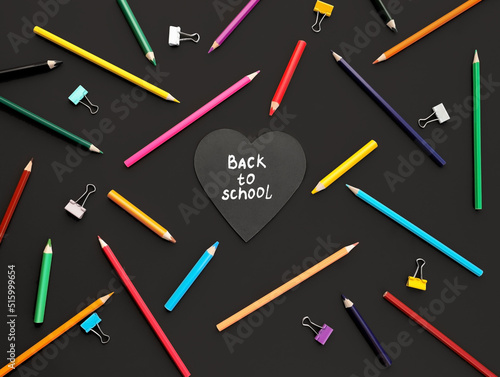 heart shaped board with back to school inscription colored pencils in chaotic order, paper clips and on black background