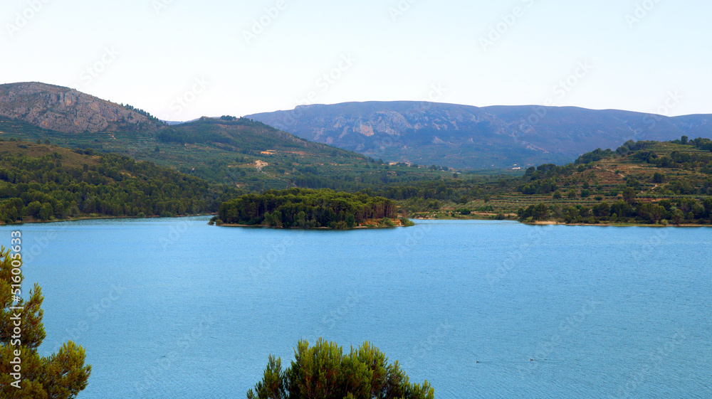 Large reservoir lake in the mountains, very blue water, pine trees on the shore, an island in the distance, mountains in the distance, beautiful landscape, Beniarres, Spain Large reservoir lake in the mountains, very blue water, pine trees on the shore, an island in the distance, mountains in the distance, beautiful landscape, Beniarres, Spain