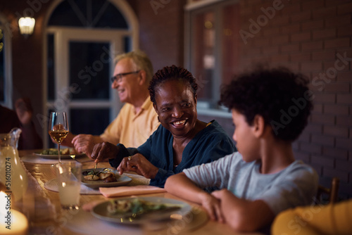 Happy black senior woman talks to her grandson during family dinner on patio.