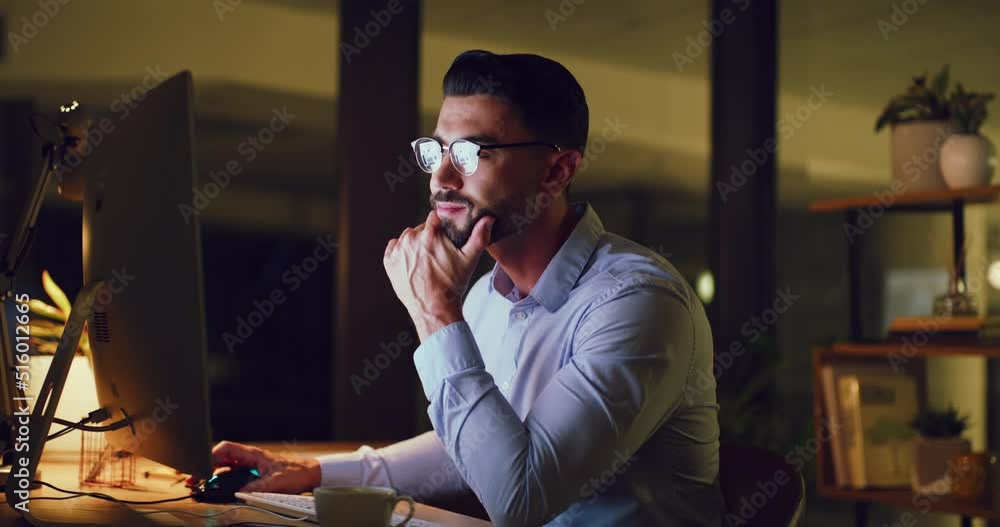 Young business man looking thoughtful while reading an email on a desktop computer and working late at night. One serious corporate male designer doing overtime to meet a deadline in a dark office