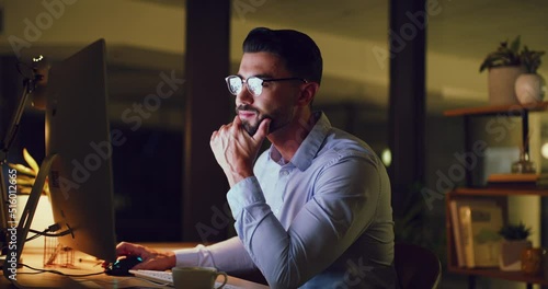 Young business man looking thoughtful while reading an email on a desktop computer and working late at night. One serious corporate male designer doing overtime to meet a deadline in a dark office photo
