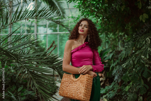 фотографія Fashionable curly woman wearing pink asymmetric ruffled top,  holding straw wicker top handle bag, posing in tropical garden