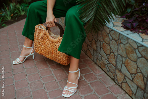 Trendy summer straw wicker top handle bag, white strap sandals in stylish female outfit. Fashion details. Copy, empty space for text photo