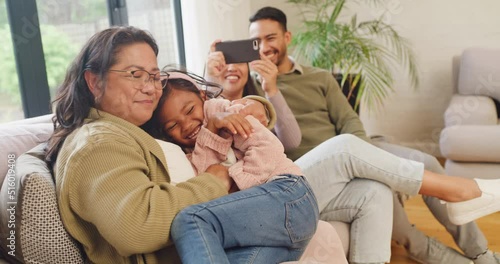 Adorable little girl playing with grandmother, laughing in home living room. Multi generation family enjoying quality time together. Smiling child being tickled while parents use a phone for photos