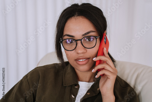 Close up of confident afro american young woman in white t-shirt and dark shirt in glasses looking at camera, holding smartphone. Student listening with attention interlocutor. Education and develop.