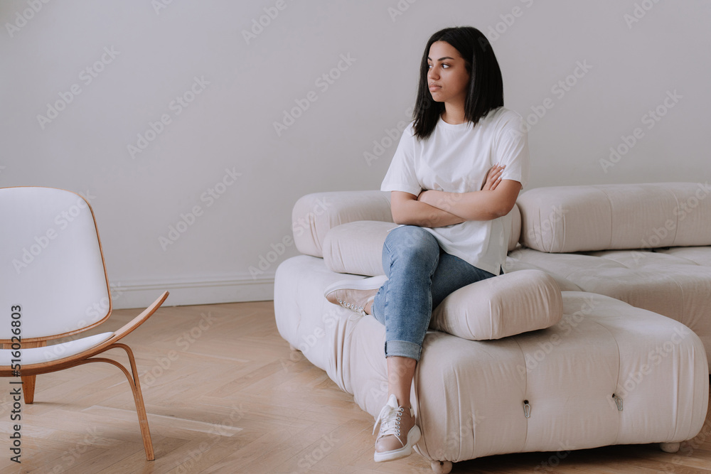 Upset afro Brazilian young woman sitting on cozy sofa at home alone. Frustrated African american girl in white t- shirt, blue jeans at home disappointed by financial troubles, divorce, rent price. Upset afro Brazilian young woman sitting on cozy sofa at home alone. Frustrated African american girl in white t- shirt, blue jeans at home disappointed by financial troubles, divorce, rent price.