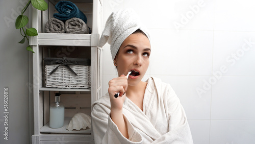Cute young woman brushing her teeth in the bathroom, wearing a bathrobe and with her hair wrapped in a towel. Daily hygiene and oral care routine.