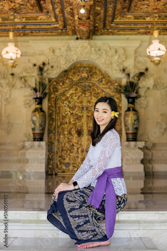 an asian woman is wearing white kebaya and batik kemben which is traditional clothes from Bali and is sitting in front of a temple wall