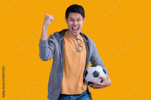 Excited asian adult man football fan in yellow t-shirt hood cheer up support favorite team in hand soccer ball hand rising up winner gesture action pose celebrate victory studio shot