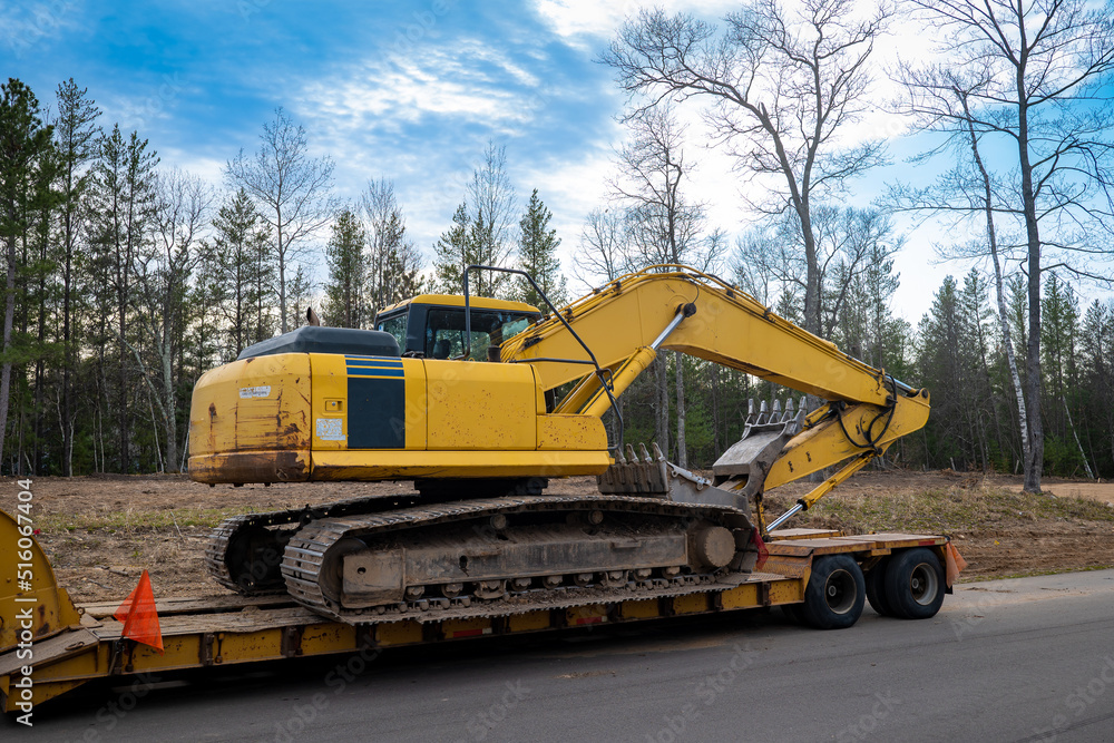 Used Yellow Skid Steer Excavator Loader on a flat bed trailer, at a construction job site, in evening light with trees in the background.