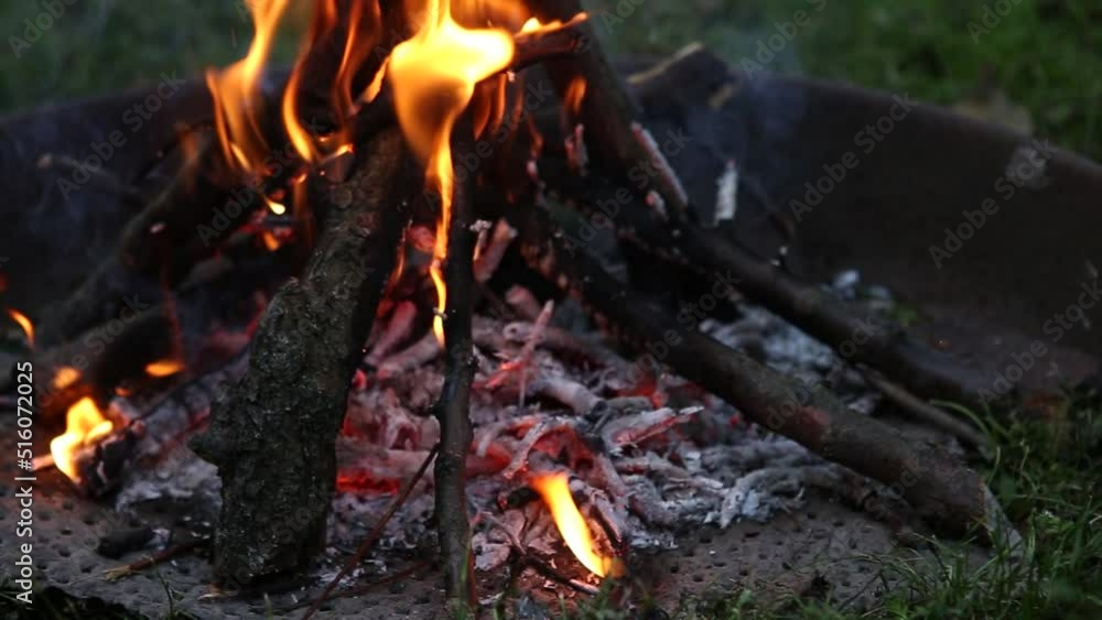 Bright flame of burning bonfire in a metal ring on the grass in evening at sunset of day in summer. Burning branches in the fire, tongues of flame in dark - closeup. Hiking. Tourism. Travel. Lifestyle Bright flame of burning bonfire in a metal ring on the grass in evening at sunset of day in summer. Burning branches in the fire, tongues of flame in dark - closeup. Hiking. Tourism. Travel. Lifestyle