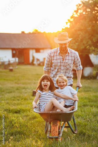 Happy little children having fun in a wheelbarrow pushing by dad in domestic garden on warm sunny day. Active outdoors games for family with kids. Spending time together on weekend at the countryside.