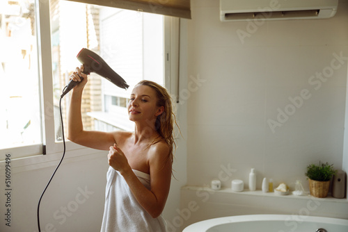Happy joyful young pretty female in towel drying hair with dryer after taking bath.