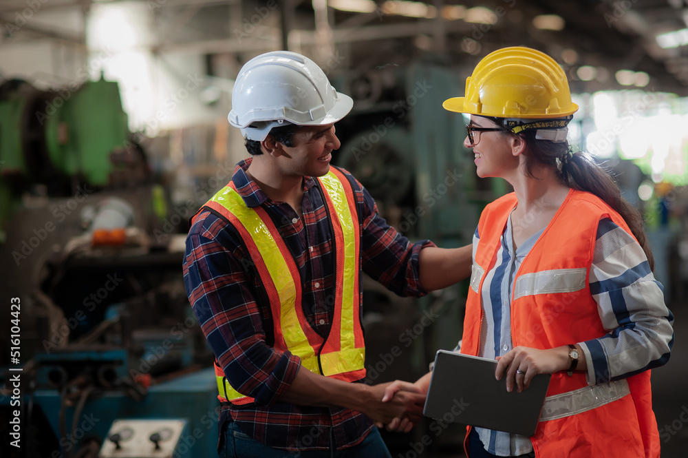 A smiling female engineer shakes hands to greet a tall, handsome, dark-skinned engineer wearing workwear safety uniform while working in an industrial factory.Teamwork and gender diversity concept. A smiling female engineer shakes hands to greet a tall, handsome, dark-skinned engineer wearing workwear safety uniform while working in an industrial factory.Teamwork and gender diversity concept.