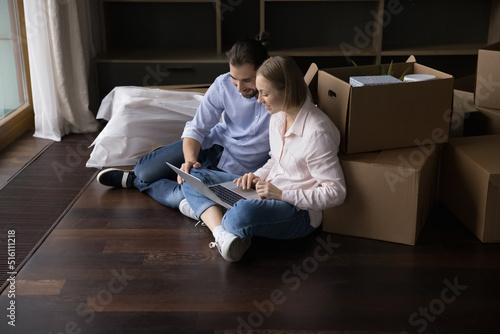 Positive millennial married couple of renters, homeowners moving into new home, sitting at heap of boxes, using laptop computer, chatting, making online order of transportation, packing service © fizkes
