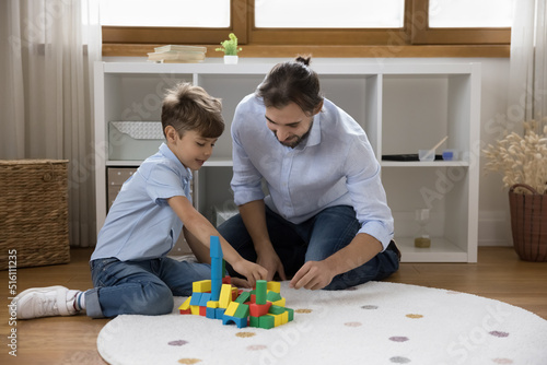 Happy engaged Caucasian young dad and son kid stacking toy tower, castle model from colorful building blocks, playing constructing game on warm carpeted floor. Family, fatherhood concept