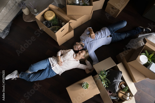 Happy peaceful couple of house buyers tired of cardboard boxes, moving, relocation, lying on floor, resting at containers with household stuff, laughing, talking. Aerial view