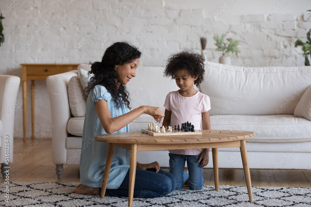Young African mother and little 5s adorable child girl play together chess in cozy living room. Caring mom develop her daughter, teach board game. Family hobby, children growth and education concept Young African mother and little 5s adorable child girl play together chess in cozy living room. Caring mom develop her daughter, teach board game. Family hobby, children growth and education concept