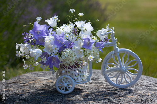A bouquet of summer white and blue bluebell flowers, lavender, hydrangea, gypsophila in a decorative bicycle on a stone in the garden, against the background of grass and lavender bushes.