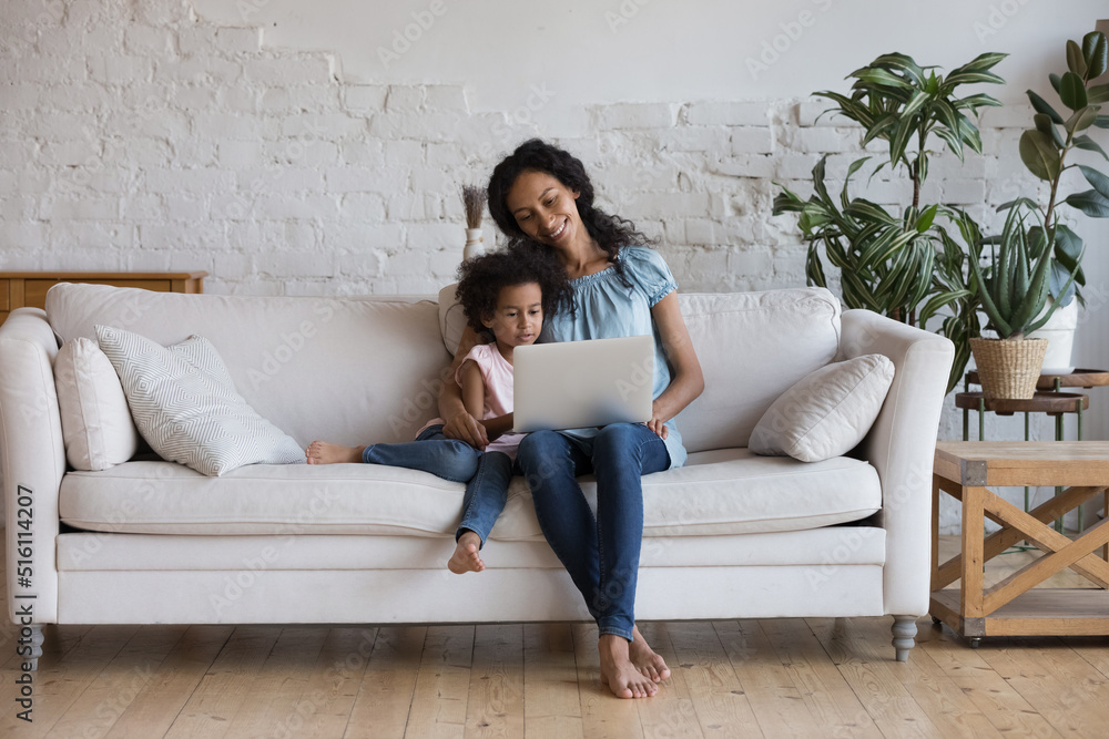 African woman and preschool cute daughter resting on sofa spend leisure on internet, watch on-line movie, buy goods using laptop. Young generation and tech usage for fun, pastime and education concept African woman and preschool cute daughter resting on sofa spend leisure on internet, watch on-line movie, buy goods using laptop. Young generation and tech usage for fun, pastime and education concept