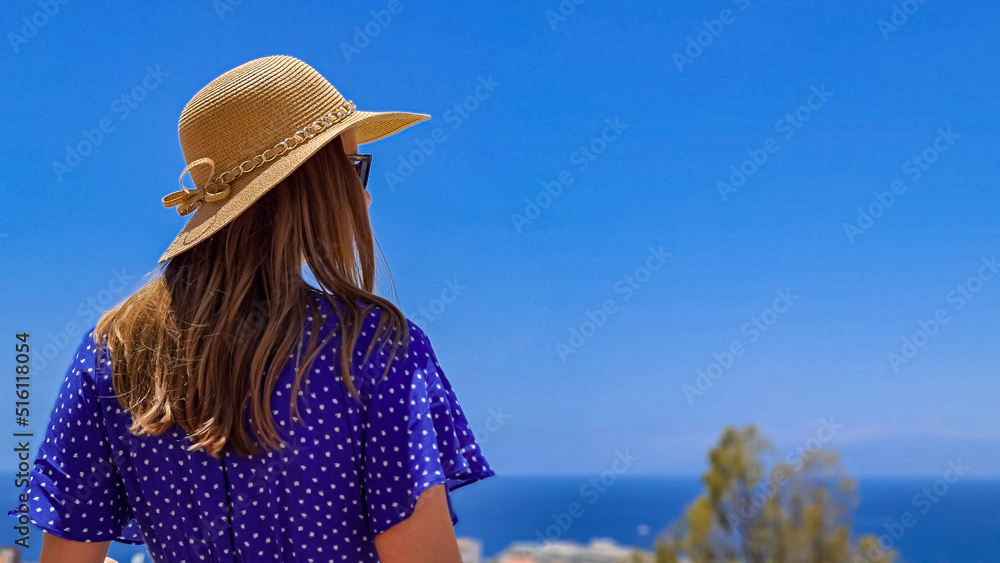 Rear view on tourist woman in summer hat and blue dress on a balcony on a luxury resort hotel on Costa Adeje, Tenerife, Canary Islands, Spain, Europe, EU. Vacation vibes. Sea view on Atlantic Ocean