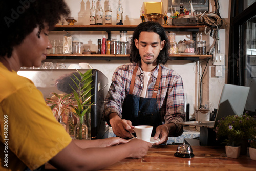 In front close-up, A young long hair Thai male cafe barista works by serving a cup of coffee to African American female customer at counter bar of coffee shop, happy service job, and SME entrepreneur.
