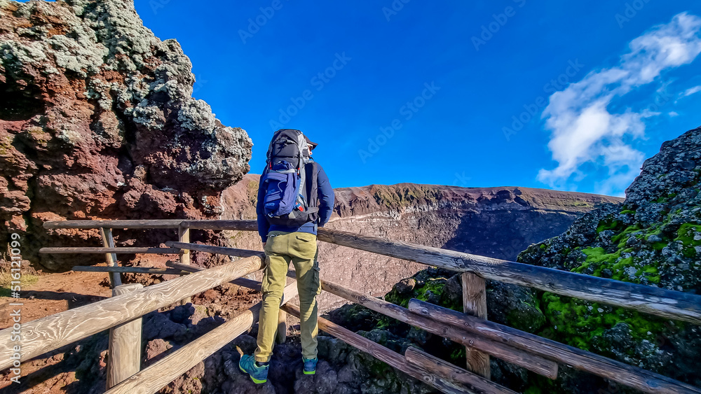 Active Man with backpack standing on fence at the edge of active volcano crater of Mount Vesuvius, Province of Naples, Campania, Italy, Europe, EU. Person enjoying the view on the mountain summit. Awe