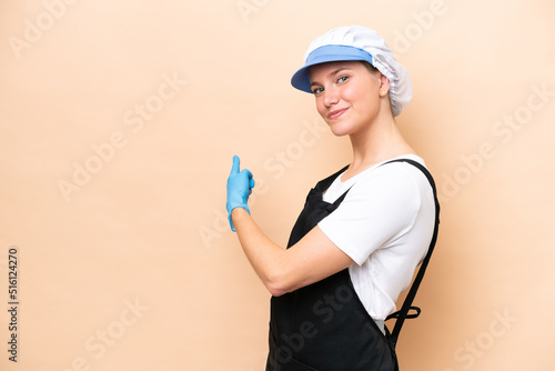 Fishmonger caucasian woman wearing an apron and holding a raw fish isolated on beige background pointing back