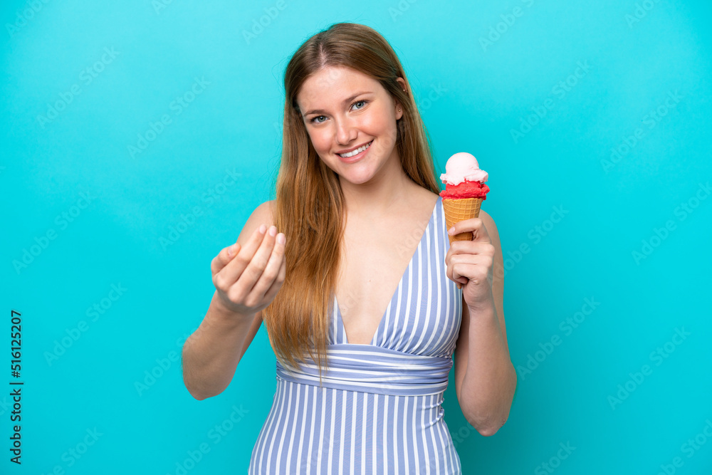 Young caucasian woman in swimsuit eating ice cream isolated on blue background inviting to come with hand. Happy that you came Young caucasian woman in swimsuit eating ice cream isolated on blue background inviting to come with hand. Happy that you came