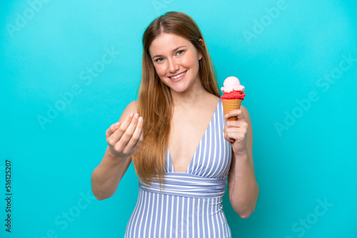 Young caucasian woman in swimsuit eating ice cream isolated on blue background inviting to come with hand. Happy that you came