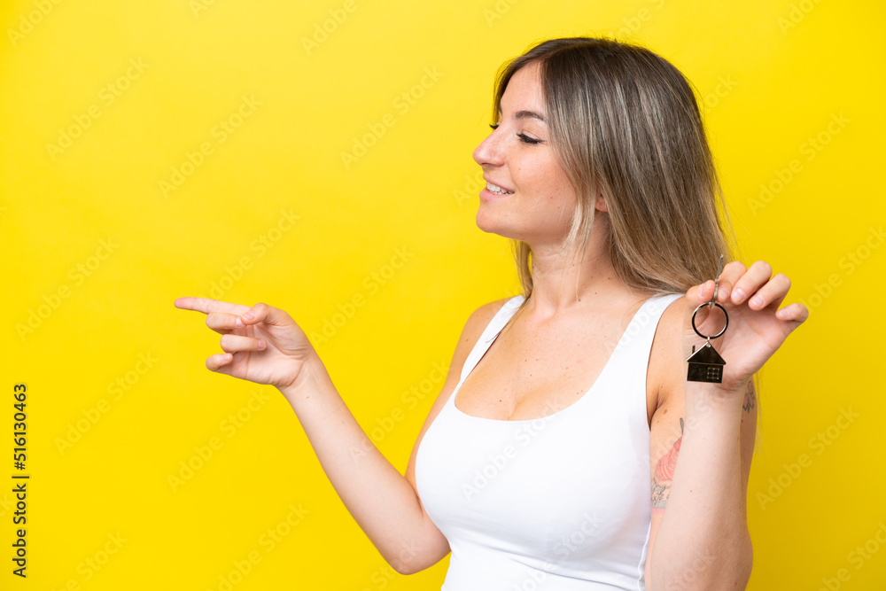 Young Rumanian woman holding home keys isolated on yellow background pointing to the side to present a product Young Rumanian woman holding home keys isolated on yellow background pointing to the side to present a product