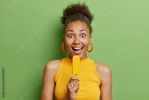 Glad surprised teenage girl with dark curly hair eats tasty frozen ice cream wears yellow t shirt and earrings enjoys cold dessert stands against green background happy about summer vacation.