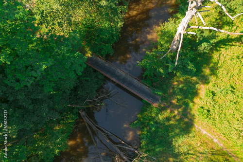 Nizinna mała rzeka. Jest słoneczny dzień. Brzegi porośnięte trawą. Nad rzeką przerzucona jest kładka dla pieszych. Jest słoneczny dzień. Zdjęcie z drona.