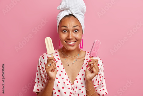 Horizontal shot of cheerful young woman holds two gelatos bites lips suggests friend to eat ice cream together wears bath towel on head isolated over pink background. Delicious summer dessert