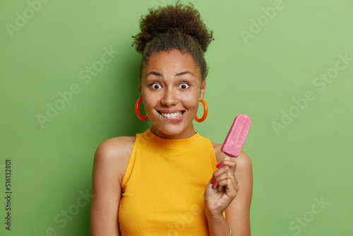 Horizontal shot of beautiful surprised woman bites lips holds appetizing ice cream with strawberry flavor enjoys sweet taste of frozen dessert dressed in yellow t shirt isolated over green background