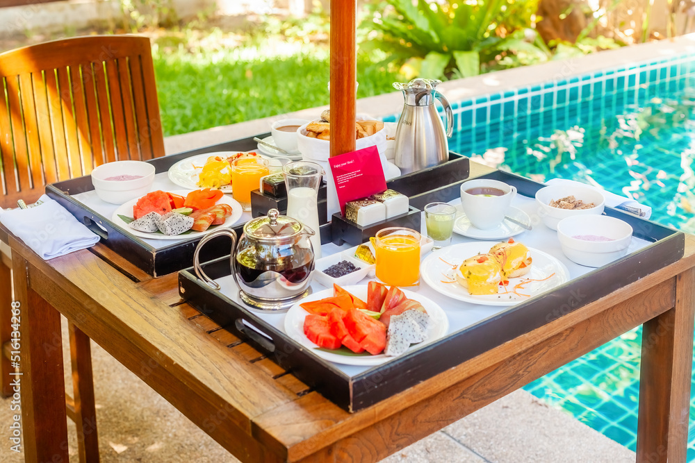 Breakfast served on villa at resort or hotel by private swimming pool. Tray with eggs benedict, fresh pastry, fruits, juice and coffee cup on poolside table. Summer travel, holidays, vacation concept.