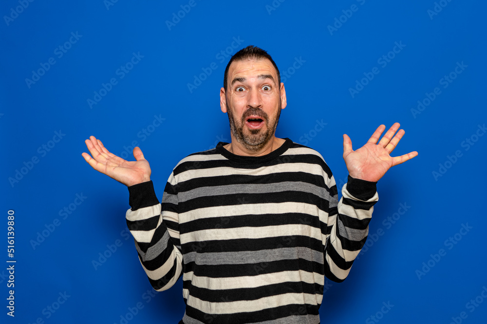 Naklejka premium Handsome bearded man in striped sweater standing over blue background scared and shocked with surprise expression, fear and excited face