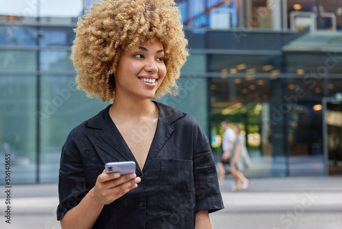 Positive curly haired beautiful woman smiles happily looks away uses smartphone while spending free time at street wears casual black t shirt messages with social follower poses at urbanity.
