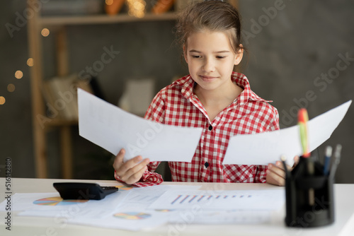 Happy little girl sitting at table reading good news in paper letter, checking internal accounts, smiling child holding paperwork, doing paperwork or studying sitting at home desk photo