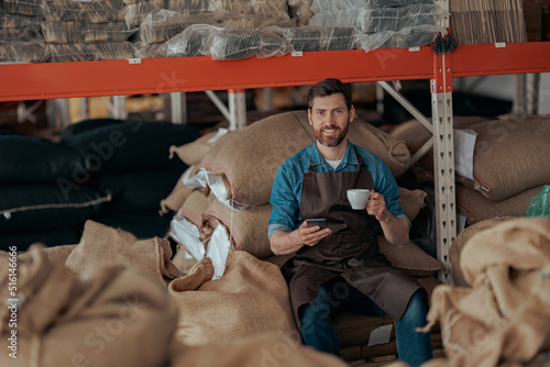 Barista sitting on the warehouse with bags of coffee beans, drinking coffee and using phone