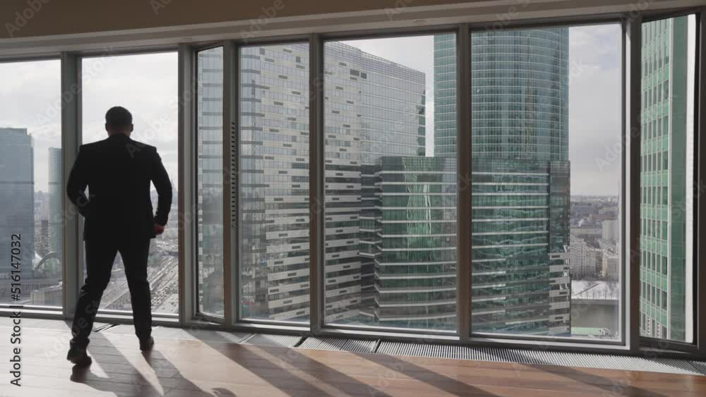 A man in a suit in the office opposite a panoramic window. A confident man approaches the window against the backdrop of skyscrapers. A man in a suit in the office opposite a panoramic window. A confident man approaches the window against the backdrop of skyscrapers.